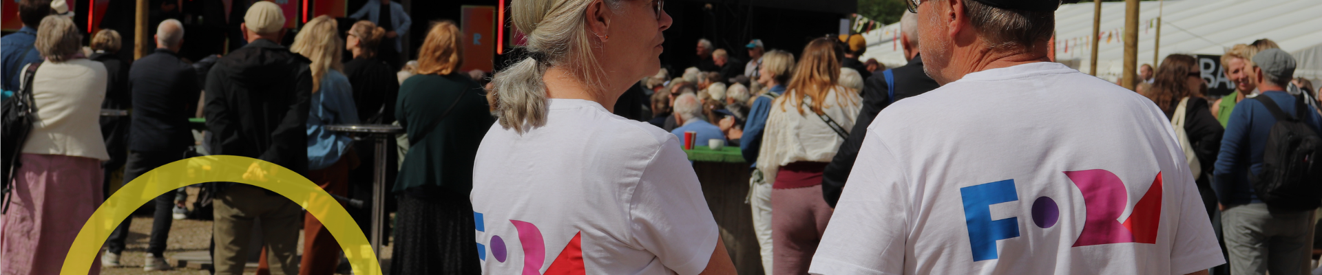 En mand og en kvinde iført en hvide t-shirts med Fora-logoer bag på, står på en festival og taler sammen.